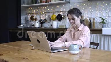 年轻女子专心致志地坐在厨房的木桌旁，用她的银色笔记本电脑和一杯